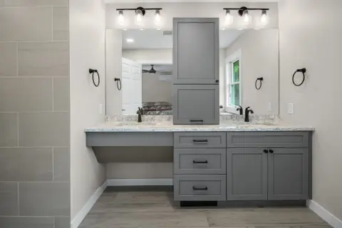 A modern, gray, freestanding vanity with dual sinks and a tall cabinet stands against a light gray wall.