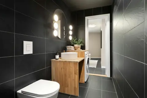 A modern bathroom with black tiles. Natural wood vanity, white sink, and toilet complement the dark tiles, showing what colour goes with black tiles in bathroom.