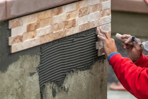 Hands expertly apply stone tile to a wall, demonstrating how to install stone tile on wall with notched trowel and adhesive.