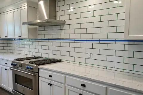 A kitchen backsplash features white subway tile with striking green grout, a modern contrast to the white cabinets and stainless steel appliances.