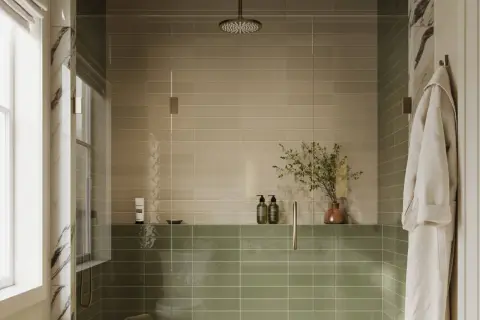 A modern shower with two-toned, low maintenance shower tile in beige and sage green. A glass enclosure, rainfall showerhead, and a plush robe complete the serene space.