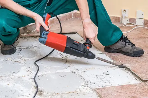 A person in green overalls uses a jackhammer to remove ceramic tile flooring, breaking up the old tiles and concrete.