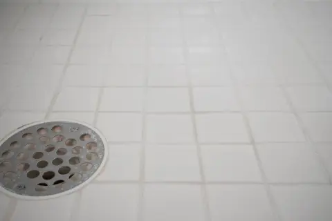 White tiled shower floor with a metal drain cover. The grout lines show signs of wear, indicating a need for tile grout repair.