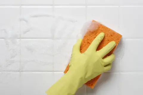 A gloved hand scrubs white bathroom tiles with a soapy orange sponge, showing the best way to clean tiles.