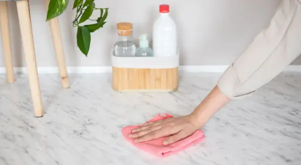 Hand wiping a marble floor with a pink cloth, with cleaning supplies in the background.