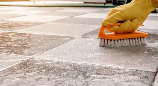 A gloved hand scrubs a tiled floor with a brush, showing the best way to clean porcelain tile.