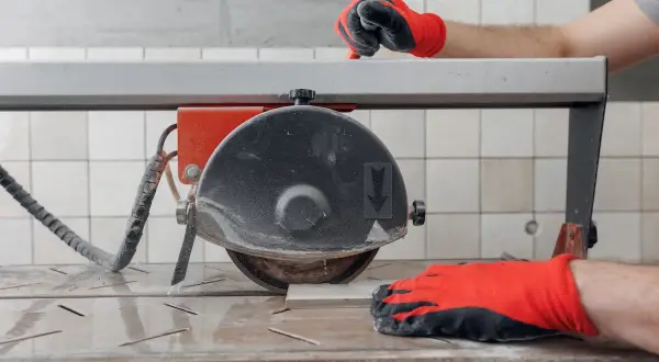 Hands in red gloves guide a tile on a wet saw, demonstrating how to use a wet saw for precise cuts.