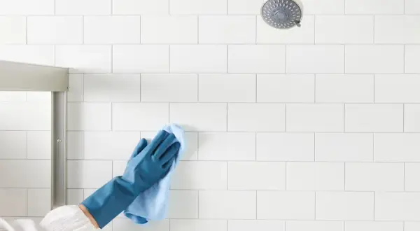 Gloved hand wipes white shower tiles, demonstrating how to clean shower tiles for a sparkling finish.