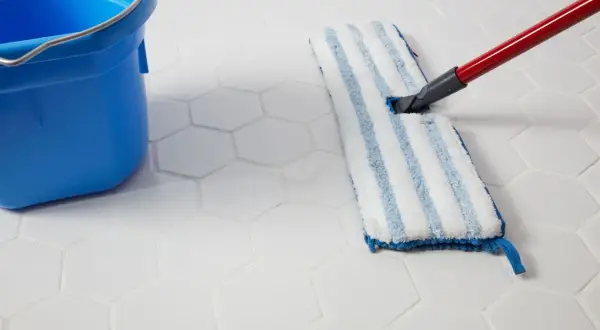 A blue bucket and a mop are used to clean white hexagonal porcelain tile floors.