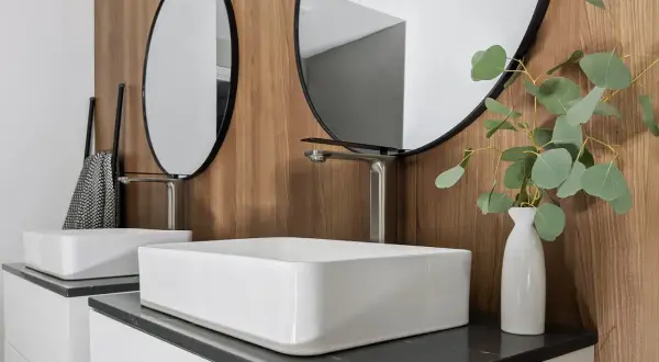 Modern bathroom with two white vessel sinks, sleek faucets, round mirrors, and a eucalyptus branch in a vase.