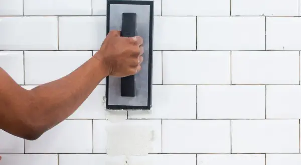 A hand applies grout to white subway tiles. The difference between sanded grout vs unsanded grout is visible in the texture.