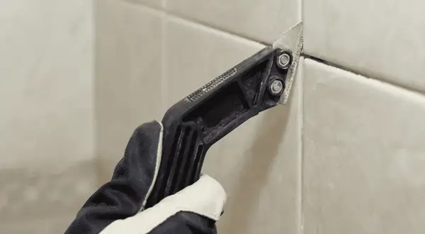 Gloved hand uses a grout removal tool to scrape away old grout, preparing for new grout over old grout.