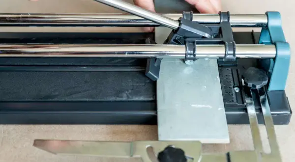 Hands guide a scoring wheel on a tile cutter, demonstrating how to cut ceramic tile by hand for a precise DIY project.