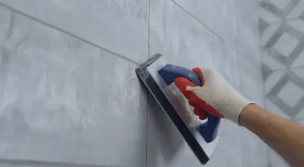 Hand in glove using a trowel for repairing grouting between light grey tiles.