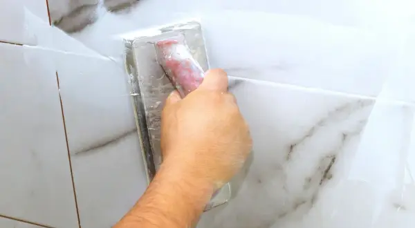 A hand uses a grout float to apply white grout to marble tiles. The grout is still wet, so it's too early to know how long does it take for grout to set.