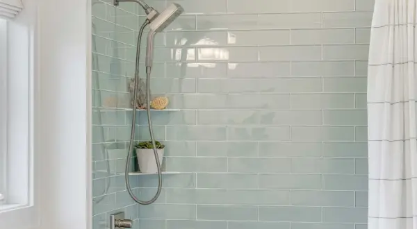Modern shower with light blue subway tiles. A shower head and hose hang next to a small plant. This is the best way to regrout tiles for a fresh look.