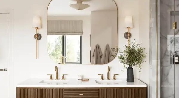 A modern bathroom vanity with dual sinks and brass fixtures. The large, rounded mirror reflects the window and wall sconces, effectively using mirrors to reflect light and brighten the space.