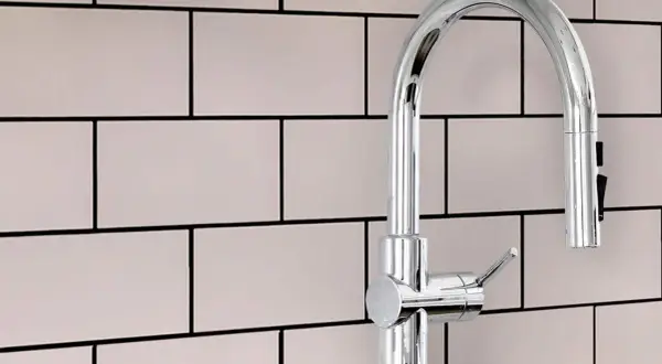 A modern chrome faucet stands against a backdrop of light pink subway tiles with smooth grout.