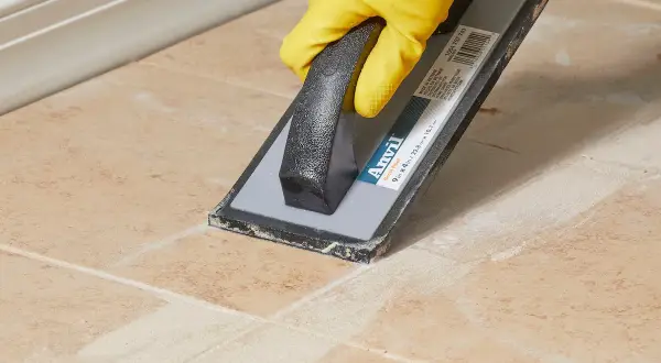 Yellow-gloved hand uses a grout float to remove old grout from tan tiles, demonstrating how to remove grout from tile.