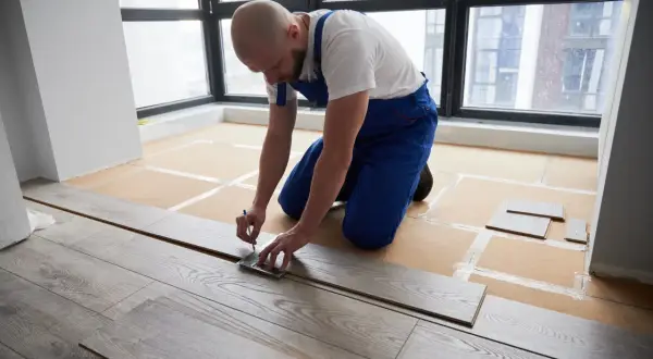 A man in blue overalls measures a laminate plank, demonstrating how to install laminate flooring.