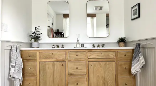 A beautiful DIY bathroom vanity with a natural wood finish, double sinks, and brass fixtures. Two framed mirrors hang above, reflecting the bright, airy space.