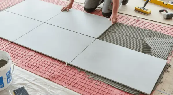 Tiling floors in a kitchen with large grey tiles and adhesive. Tools and a bucket of grout are nearby.