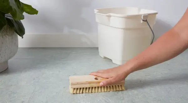 A hand scrubs a linoleum floor with a brush, with a bucket nearby. This shows how to clean linoleum floors effectively.