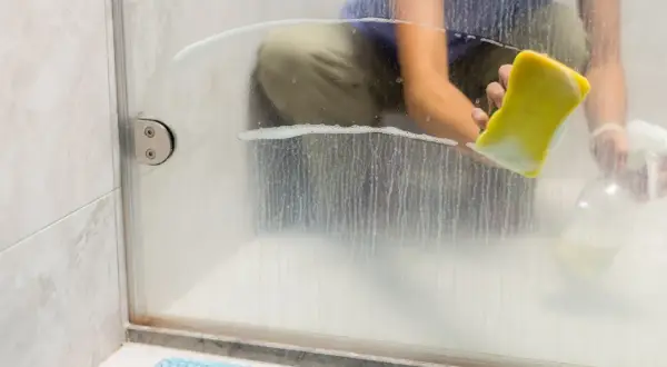 A person demonstrates how to clean shower glass with a yellow sponge and spray bottle, tackling soap scum and water spots for a sparkling finish.