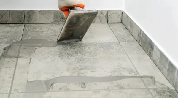 Gloved hand applies grout to tiled floor. Wondering how long for grout to dry? This process is key to a finished look.