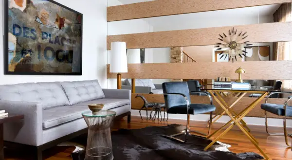 A large wall mirror with wood accents offers a stylish way to decorate around it. A grey sofa, abstract art, and a glass desk complete the modern living space.