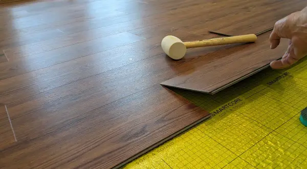 A hand is installing a dark wood-look LVT plank flooring over yellow underlayment. A rubber mallet rests nearby.