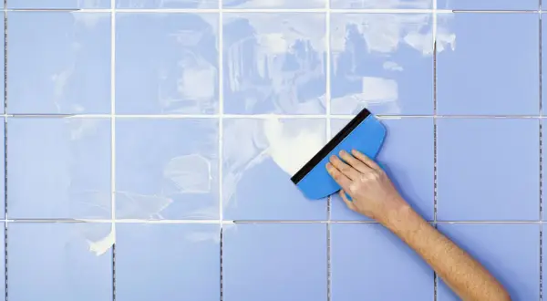 A hand uses a blue squeegee to spread white grout on light blue tiles, demonstrating how to clean tile grout.