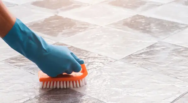 Gloved hand scrubs tile grout with an orange brush, demonstrating how to clean grout on a tile floor.