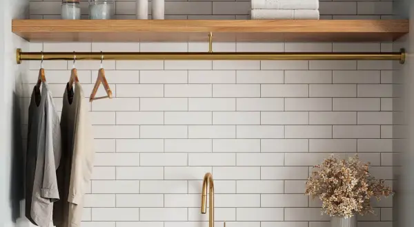 A laundry room with white tile beige grout walls, a wooden shelf, and a brass rod holding clothes.