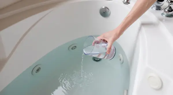A hand pours liquid from a glass into a jetted bathtub, part of a process for how to clean bathtub.