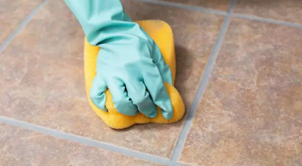 A gloved hand scrubs unglazed ceramic tile with a yellow sponge, demonstrating how to clean the porous surface.
