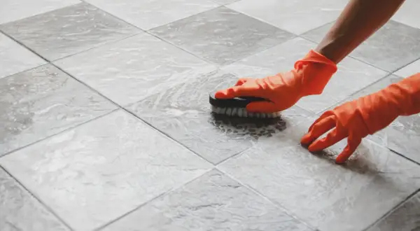 Hands in orange gloves scrub a tiled floor. The person is working to polish porcelain tile, making it shine.