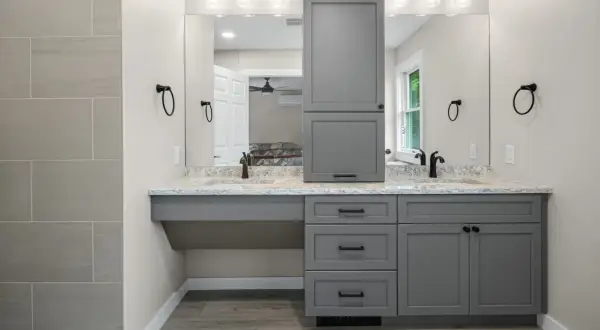 A modern, gray, freestanding vanity with dual sinks and a tall cabinet stands against a light gray wall.