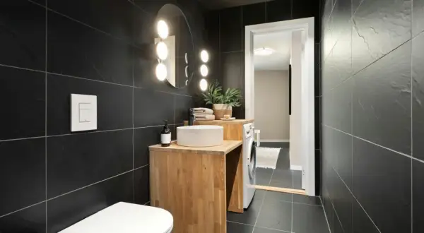 A modern bathroom with black tiles. Natural wood vanity, white sink, and toilet complement the dark tiles, showing what colour goes with black tiles in bathroom.