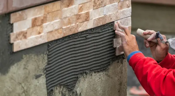 Hands expertly apply stone tile to a wall, demonstrating how to install stone tile on wall with notched trowel and adhesive.