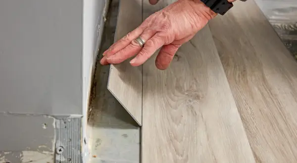 Hands installing light wood-look vinyl plank flooring. The question of how long does it take to install vinyl plank flooring depends on room size and prep.