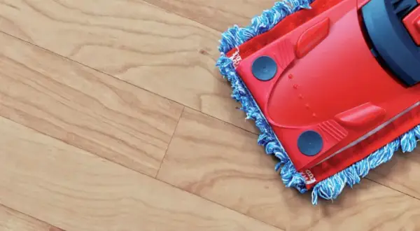 A red mop with blue fringe is cleaning a light wood-look vinyl floor, leaving it spotless.