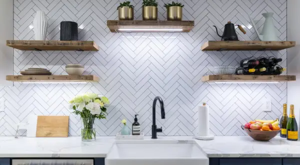 A modern kitchen with a white chevron tile backsplash, rustic wood shelves, and a farmhouse sink. The chevron tile out of style? Not in this chic design!