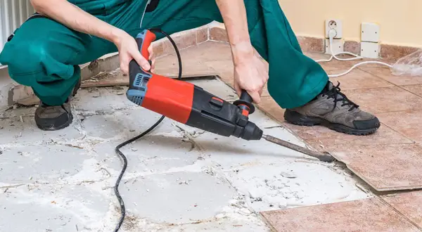 A person in green overalls uses a jackhammer to remove ceramic tile flooring, breaking up the old tiles and concrete.
