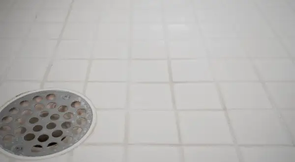 White tiled shower floor with a metal drain cover. The grout lines show signs of wear, indicating a need for tile grout repair.