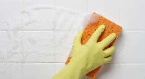 A gloved hand scrubs white bathroom tiles with a soapy orange sponge, showing the best way to clean tiles.
