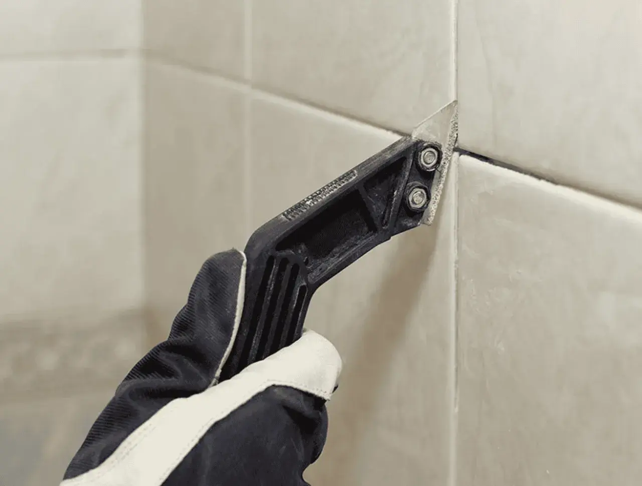 Gloved hand uses a grout removal tool to scrape away old grout, preparing for new grout over old grout.
