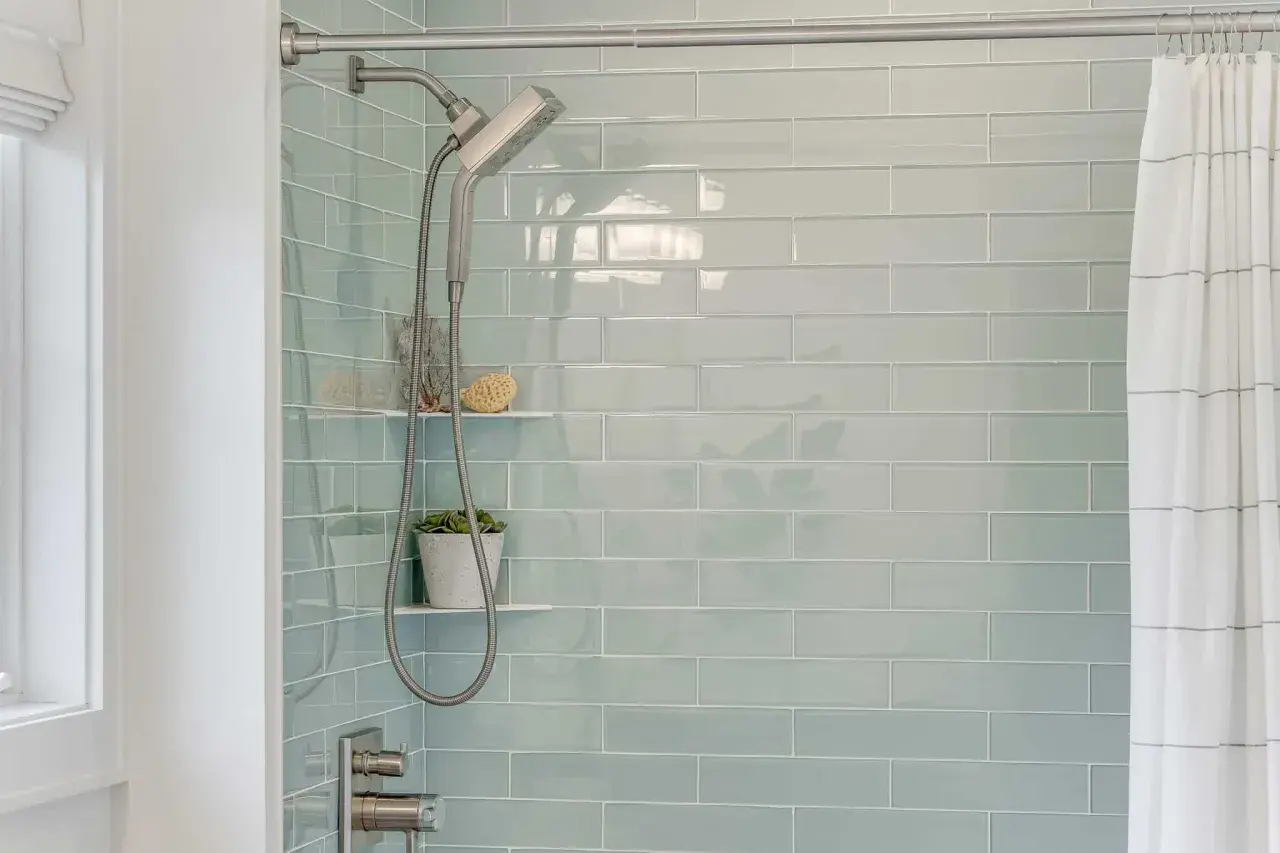 Modern shower with light blue subway tiles. A shower head and hose hang next to a small plant. This is the best way to regrout tiles for a fresh look.