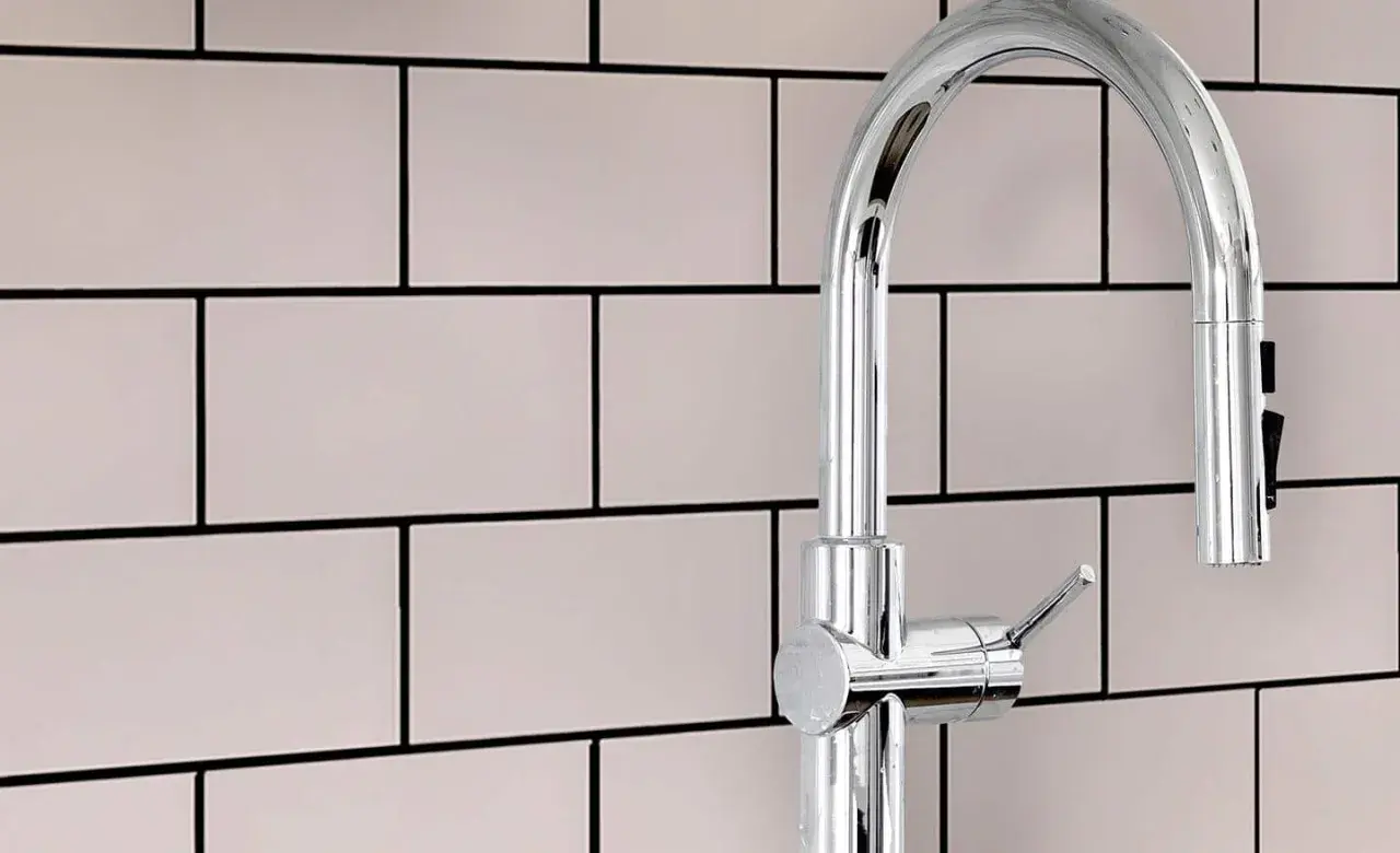 A modern chrome faucet stands against a backdrop of light pink subway tiles with smooth grout.