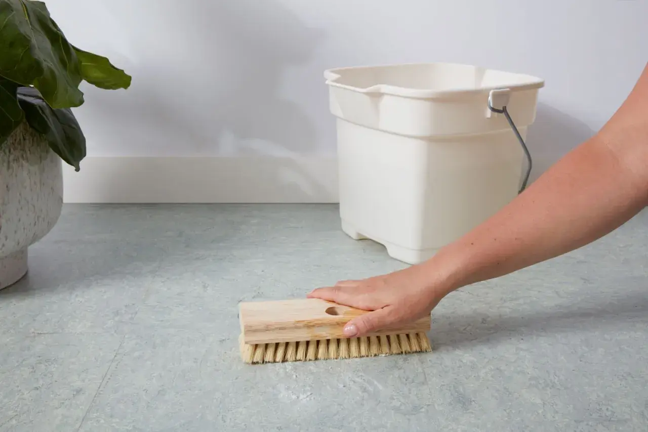 A hand scrubs a linoleum floor with a brush, with a bucket nearby. This shows how to clean linoleum floors effectively.